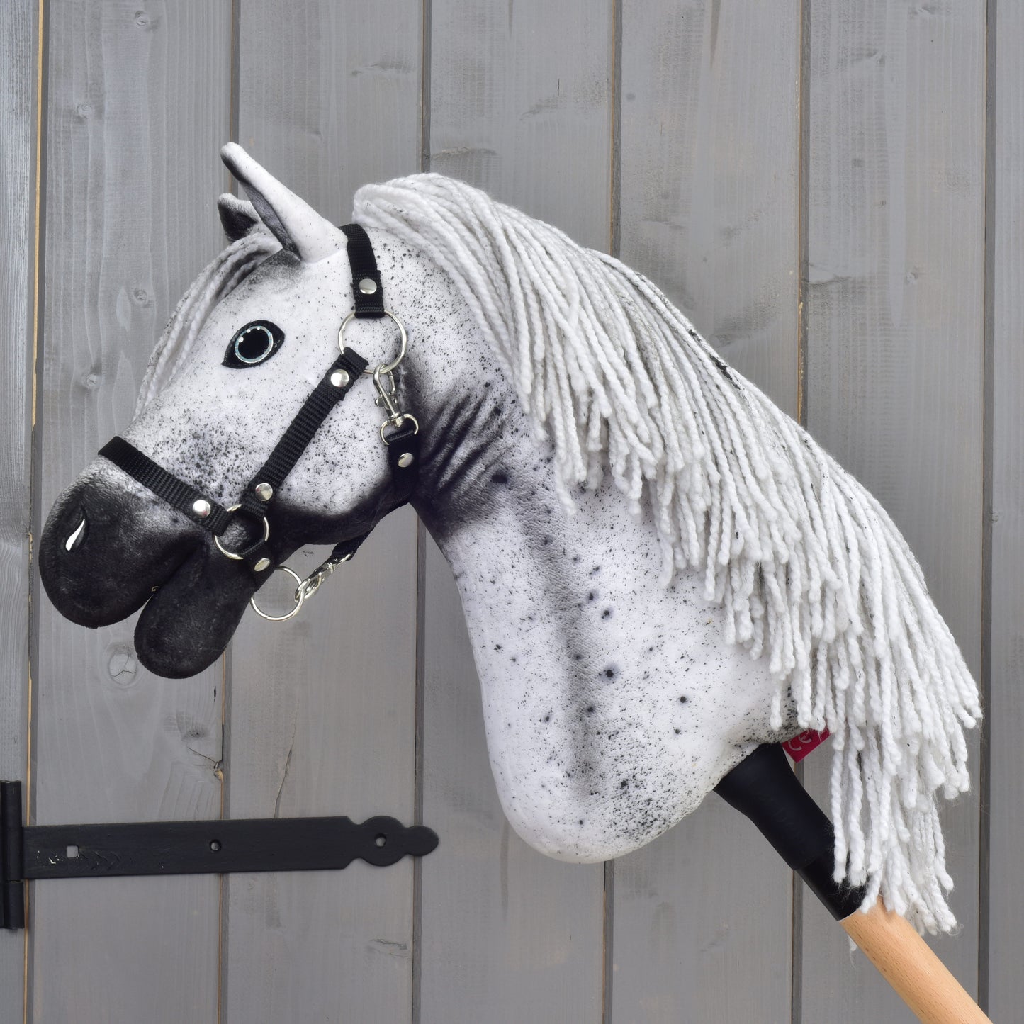 Hobby Horse Naivy met zwart halster - maat S van Hobby Horse LarDen, een witte en grijze Hobby Horse met manen van garen, gemonteerd op een houten stok tegen een grijze houten wand.