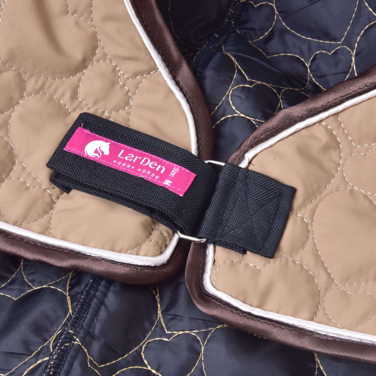 Close-up van de lichtbruine en bruine gewatteerde Halsbeschermer Bonanza van Hobby Horse LarDen, met een zwarte riem met veilige sluitingen, een metalen ring en een roze label met de merknaam en een paardenlogo.