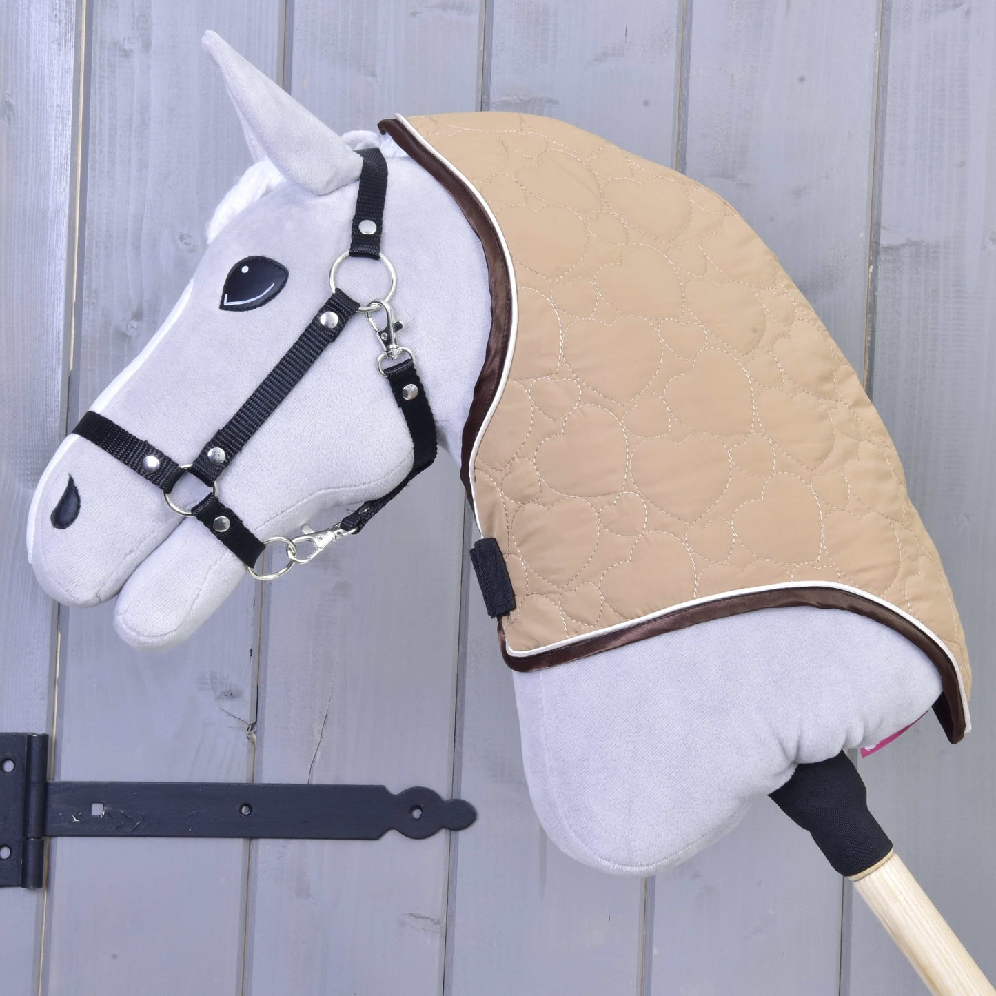 Een close-up van de Halsbeschermer Bonanza van Hobby Horse LarDen: een grijze Hobby Horse met zwart hoofdstel en lichtbruine gewatteerde deken met veilige sluitingen, gemonteerd op een houten stok voor een grijze houten wand.