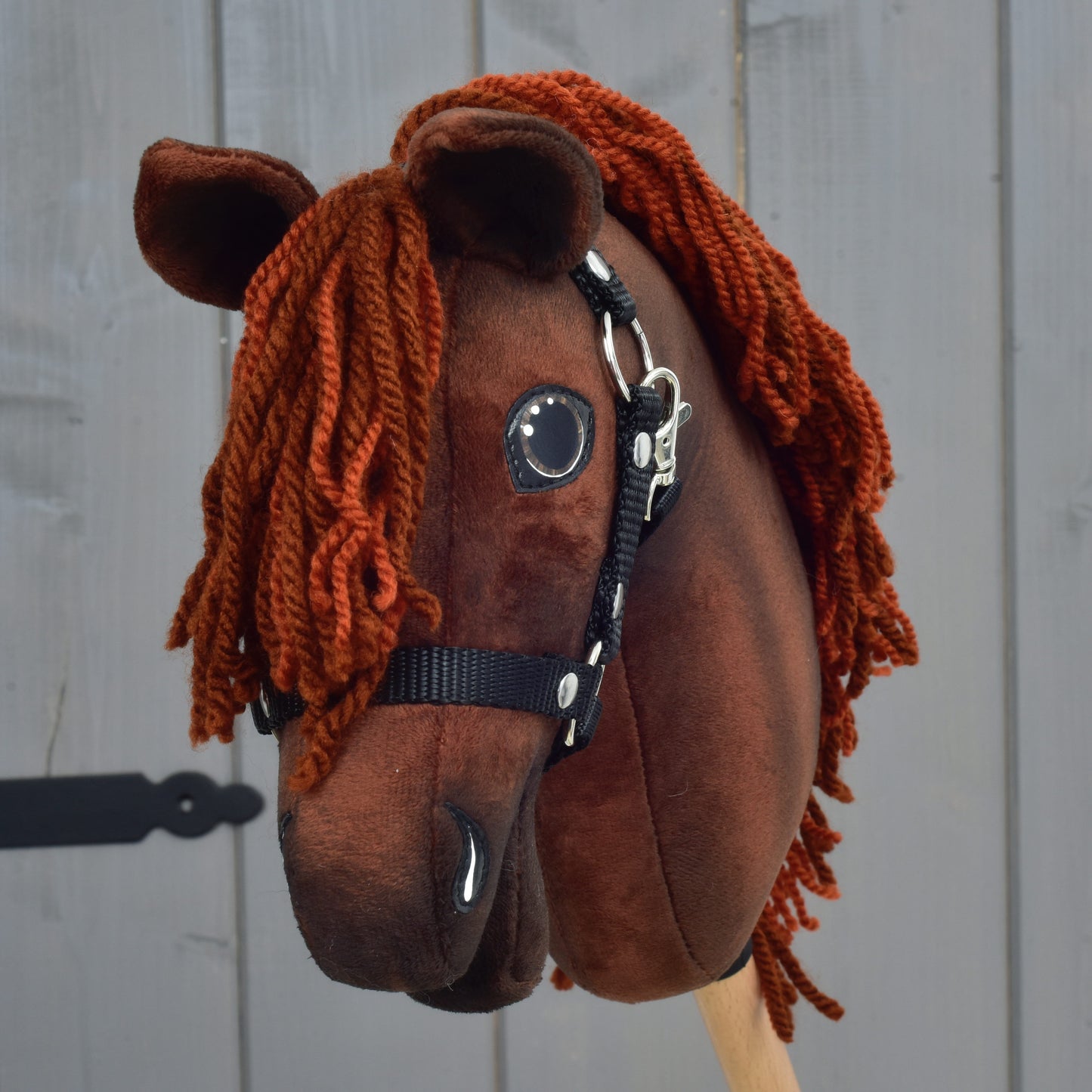 A brown fabric Hobby Horse Rebel with Black Halter - size XS from Hobby Horse LarDen, designed to resemble a Hanoverian stallion with an orange yarn mane, is displayed in front of a gray wooden wall.