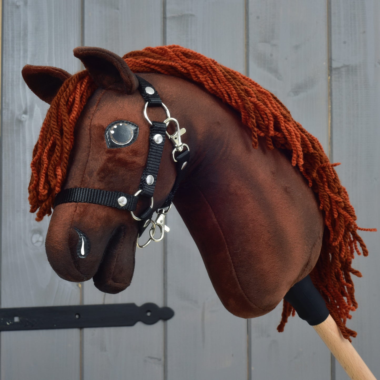 A Hobby Horse Rebel with Black Halter - size XS by Hobby Horse LarDen, featuring a brown Hanoverian design with a red yarn mane, is mounted on a wooden stick and displayed in front of a gray wooden wall.