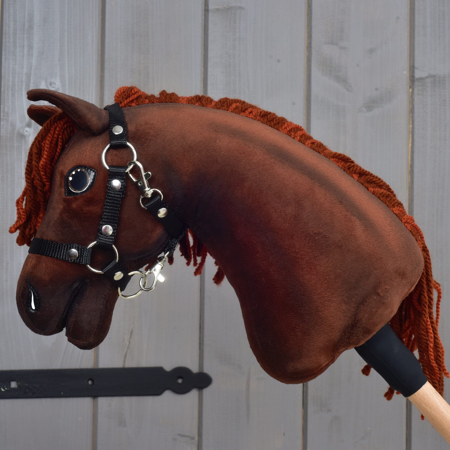 A Hobby Horse LarDen "Rebel" with a black halter (size XS), featuring brown plush fabric and an orange yarn mane, is mounted on a wooden stick in front of a gray wooden wall.
