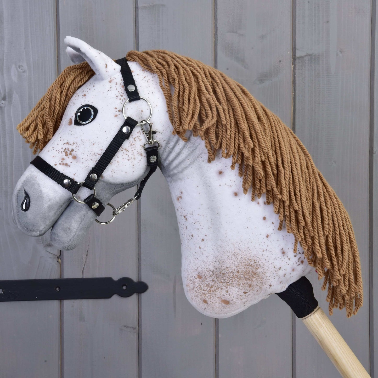 Close-up van Hobby Horse Rufus van Hobby Horse LarDen met een wit gezicht, bruine manen van garen, zwart halster en bruine spikkels, tegen een achtergrond van houten panelen.