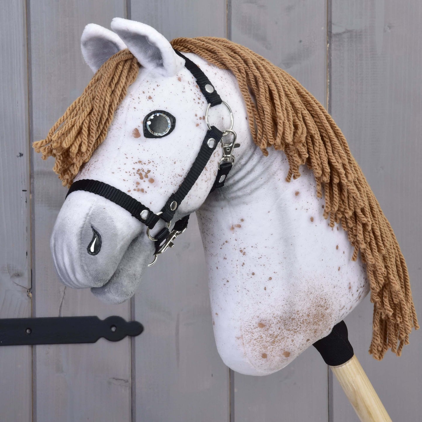 Hobby Horse Rufus met zwart halster van Hobby Horse LarDen heeft een handgemaakt wit gespikkeld stoffen hoofd, bruine manen van garen, knoopogen en een zwart halster op een houten stok, weergegeven tegen een grijze houten achtergrond.