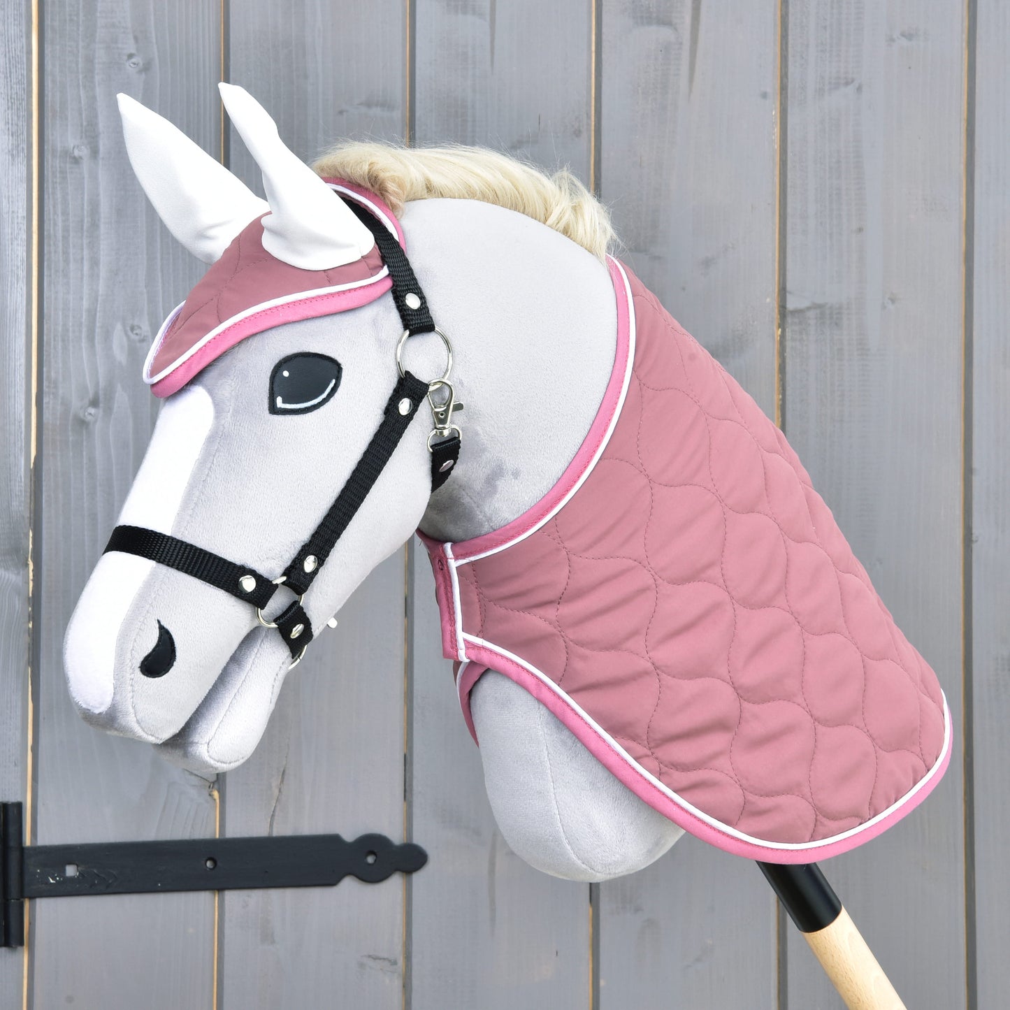 The Hobby Horse LarDen "Set Dusty Rose" features a gray hobby horse with a white mane, black bridle, ear bonnet, quilted pink blanket, and a black stick handle, displayed against a wooden wall for added charm.