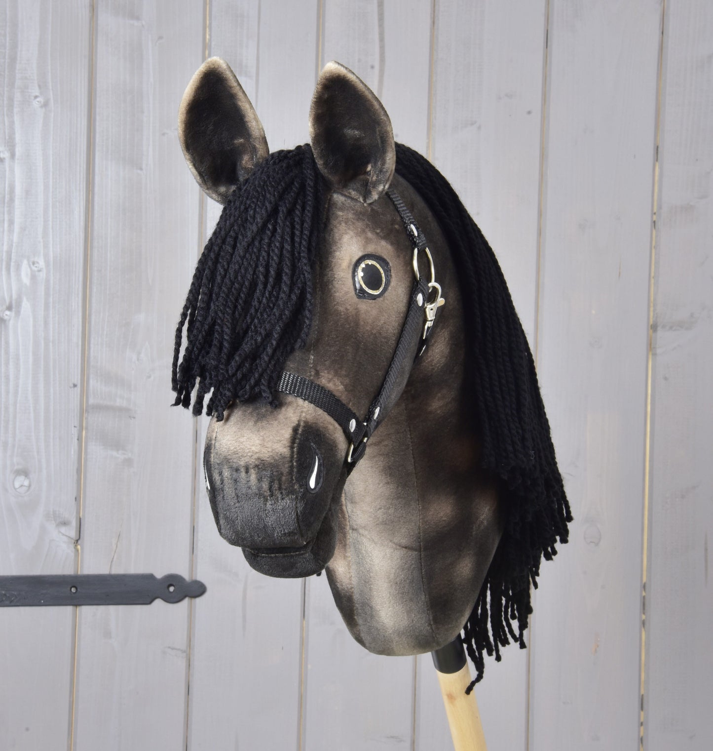 Hobby Horse Stormy with Black Halter by Hobby Horse LarDen: a dapple grey plush horse head with a long black yarn mane, soulful eye and black halter, mounted on a wooden stick against a light wooden wall.