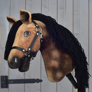 Hobby Horse Stormy met zwart halster van Hobby Horse LarDen, met een bruin pluche Hannoveraans ontwerp, zwarte garenmanen, zwart halster en witte aftekeningen, gemonteerd op een stok tegen een grijze houten achtergrond.