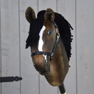 Hobby Horse Sundance met zwart halster van Hobby Horse LarDen met een bruine vacht, witte bles, zwarte manen van garen en zwart halster, lijkend op een levendige volbloedhengst, afgebeeld tegen een lichtgrijze achtergrond van houten planken.
