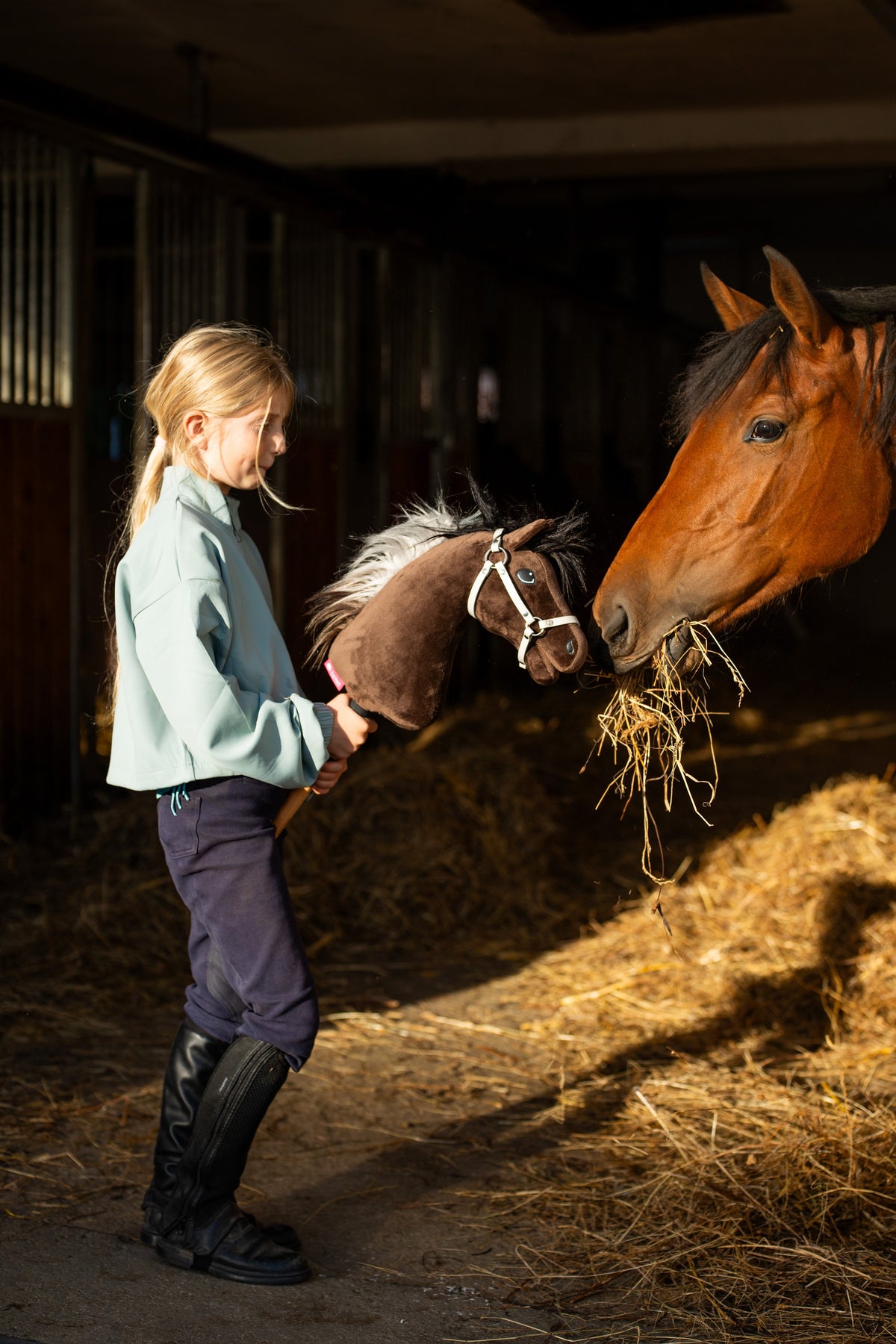 Meisje in mintgroen shirt en rijlaarzen in een stal met Hobby Horse Alfredo in witte halster naast een bruin paard dat hooi eet