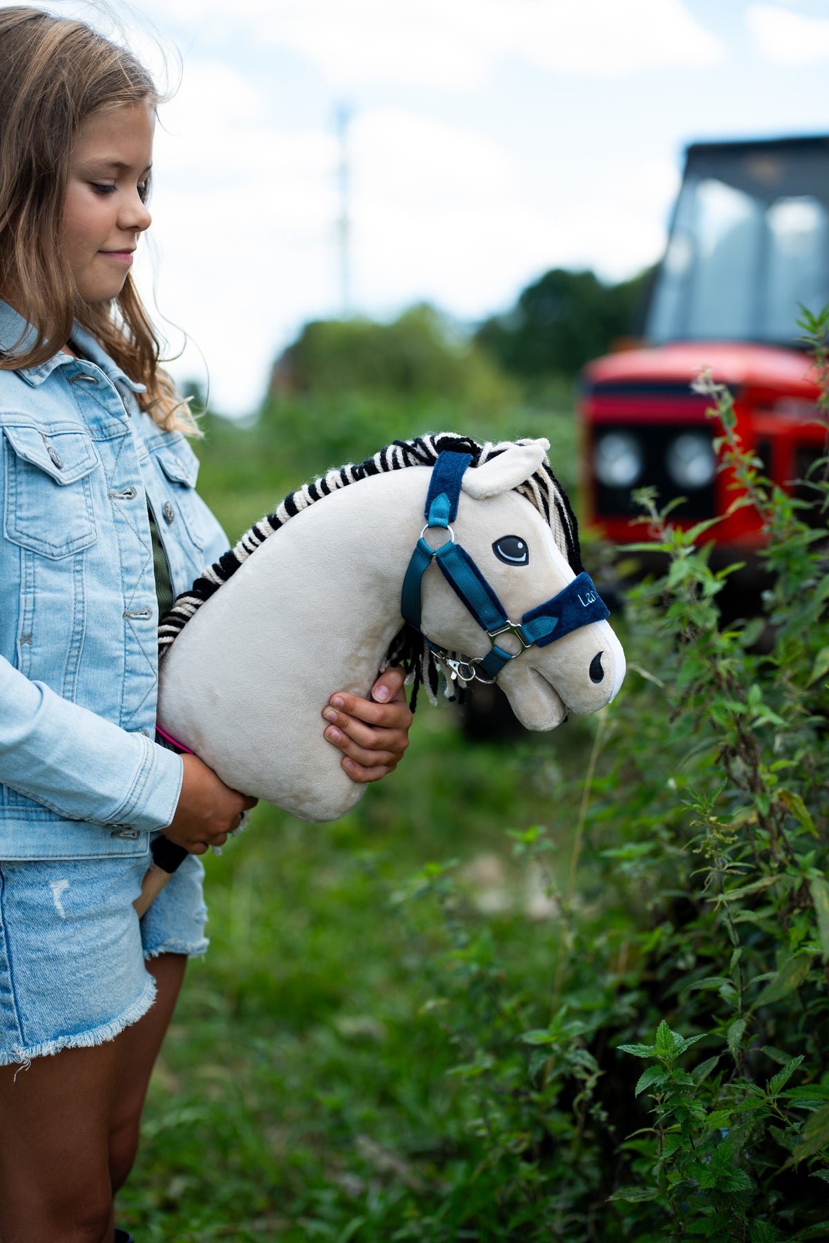 Lifestylefoto: een meisje in een spijkerjasje houdt de Hobby Horse Aurora met zwart Halster van Hobby Horse LarDen naast een heg vast, met een rode tractor vaag op de achtergrond - crèmewitte pluchen kop, zwart-wit gestreepte garenmane - hier getoond met het met bont gevoerde LarDen Blue Halster