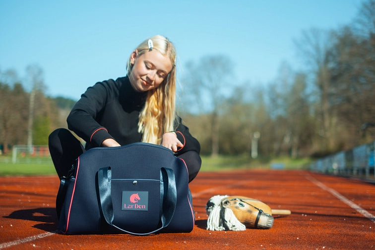 Een jonge vrouw met lang blond haar zit op een atletiekbaan en kijkt in een marineblauwe sporttas met de naam LarDen. Een hobby horse ligt op de grond naast haar en hint naar haar passie voor Hobby Horsing. Bomen en baanmarkeringen zijn zichtbaar op de achtergrond.