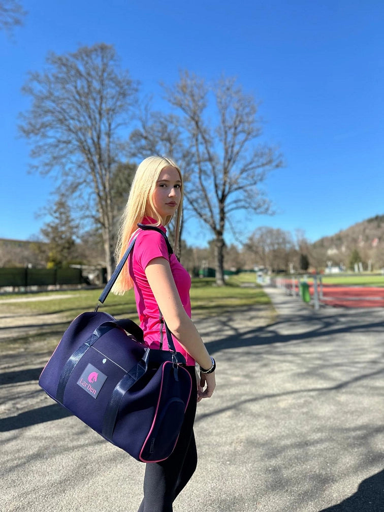 Een jonge vrouw met lang blond haar, in een roze sportshirt en met een donkerblauwe sporttas, staat buiten op een zonnige dag met haar Hobby Horse, klaar voor een leuke dag Hobby Horsing onder de heldere blauwe lucht en tussen de groene bomen.