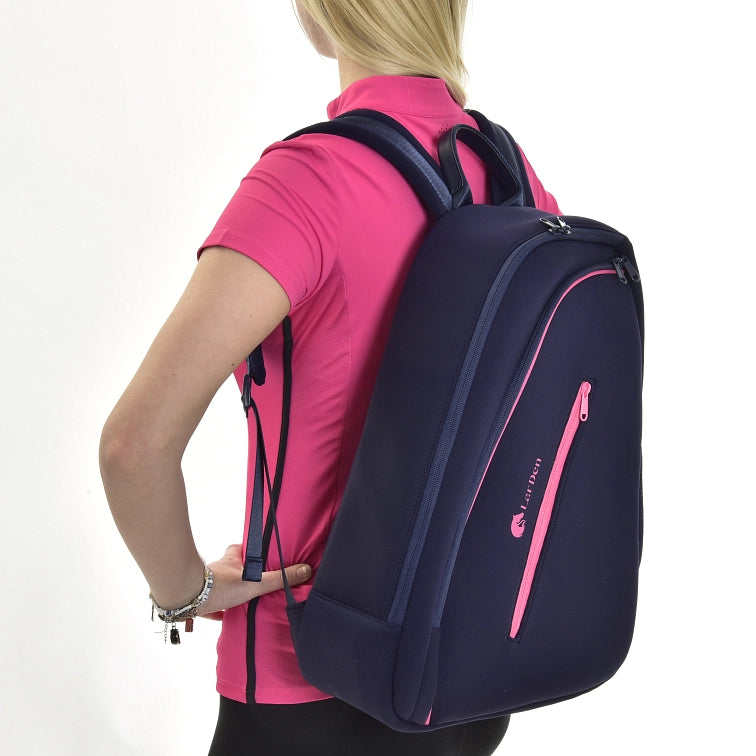 Een persoon met blond haar in een felroze shirt draagt een grote donkerblauwe rugzak met roze ritsen en accenten en houdt een Hobby Horse vast, tegen een effen witte achtergrond.