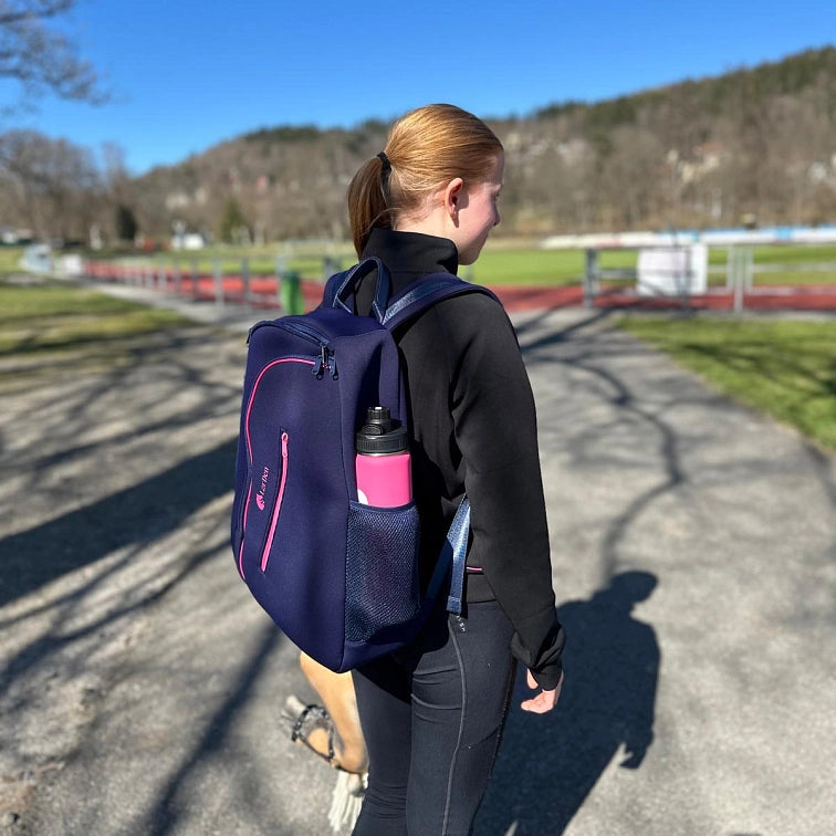 Een jonge vrouw met rood haar in een paardenstaart loopt buiten op een zonnige dag, draagt zwarte sportkleding en een donkerblauwe rugzak met een roze waterfles. Bomen en een atletiekbaan op de achtergrond suggereren dat ze op weg is naar of terugkomt van haar favoriete Hobby Horsing-activiteit.