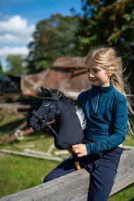 Lifestylefoto: een blond meisje in een petrolkleurige top zit op een houten reling op een zonnig erf en houdt de Hobby Horse Bond met zwart Halster van Hobby Horse LarDen vast - zwarte pluchen kop, donkerbruine garenmane - hier getoond met het versierde bitloze hoofdstel