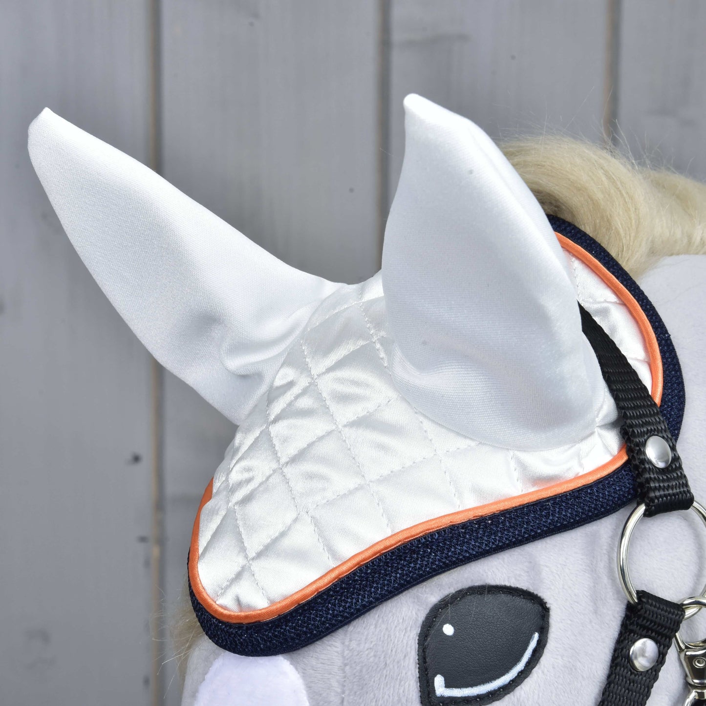 Close-up van Oornetje Speedy van Hobby Horse LarDen in wit met oranje bies, weergegeven op een pluche Hobby Horse met het linkeroog en hoofdstel zichtbaar — stijlvolle vliegenbescherming tegen een houten achtergrond.