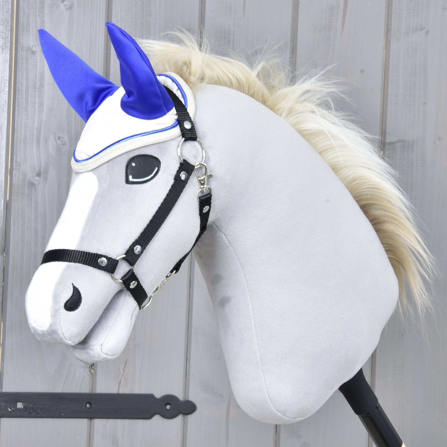 Hobby Horse LarDen met een pluchen lichtgrijze kop, witte snuit, zwart hoofdstel en crèmekleurige manen, voorzien van het Oornetje Crème Parel voor bescherming tegen insecten. Het is gemonteerd op een stok en staat tegen een houten achtergrond.