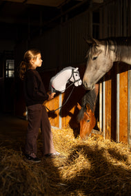 Donkerharig meisje in een zwart sweatshirt in de stalgang met Hobby Horse Caesar met zwart Halster van Hobby Horse LarDen, grijze pluchen kop met zwart-witte garenmane, terwijl een grijs paard en een bruin paard uit hun boxen kijken, met het versierde Complete bitloze hoofdstel