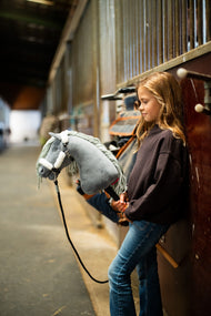 Lifestylefoto: een meisje in een donkerbruine sweater leunt tegen de stallen in een stalgang en houdt de Hobby Horse Calypso met zwart Halster van Hobby Horse LarDen vast - grijze pluchen kop, lichtgrijze garenmane - hier getoond met het met bont gevoerde Teddy Classic Halster en het zwarte Halstertouw