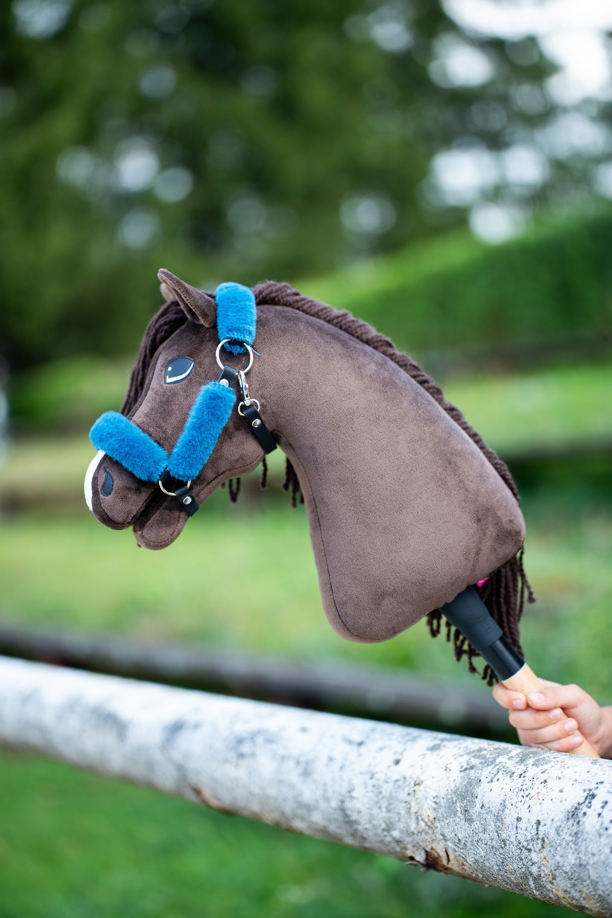 Detailfoto: de kop van de Hobby Horse Chianti met zwart Halster van Hobby Horse LarDen rust over een witte paddockreling - donkerbruin pluche, donkere garenmane en voorpluk - hier getoond met het met bont gevoerde Teddy Petrol Halster in helder blauw