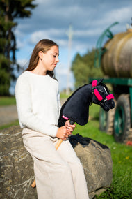 Lifestylefoto: een meisje in een crèmekleurige trui zit op een rotsblok bij een weidepad en houdt de Hobby Horse Cloony met zwart Halster van Hobby Horse LarDen vast - zwarte pluchen kop, witte bles, zwarte garenmane - hier getoond met het zwarte kunstleren Halster met volledig roze bont