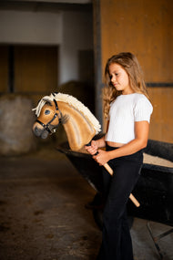 Lifestylefoto: een meisje in een witte top staat in een stal en houdt de Hobby Horse Colby met zwart Halster van Hobby Horse LarDen vast - palomino pluchen kop, dikke crèmekleurige garenmane, zwart Halster met zilveren beslag