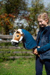 Lifestylefoto: een meisje in een donkerblauw gilet houdt de Hobby Horse Connie met zwart Halster van Hobby Horse LarDen vast, gekleed in een petrolblauwe Staldeken, met herfstbessen erachter - voskleurige pluchen kop, crèmekleurige garenmane - hier getoond met het met bont gevoerde Teddy Classic Halster en de Winter Sky Staldeken