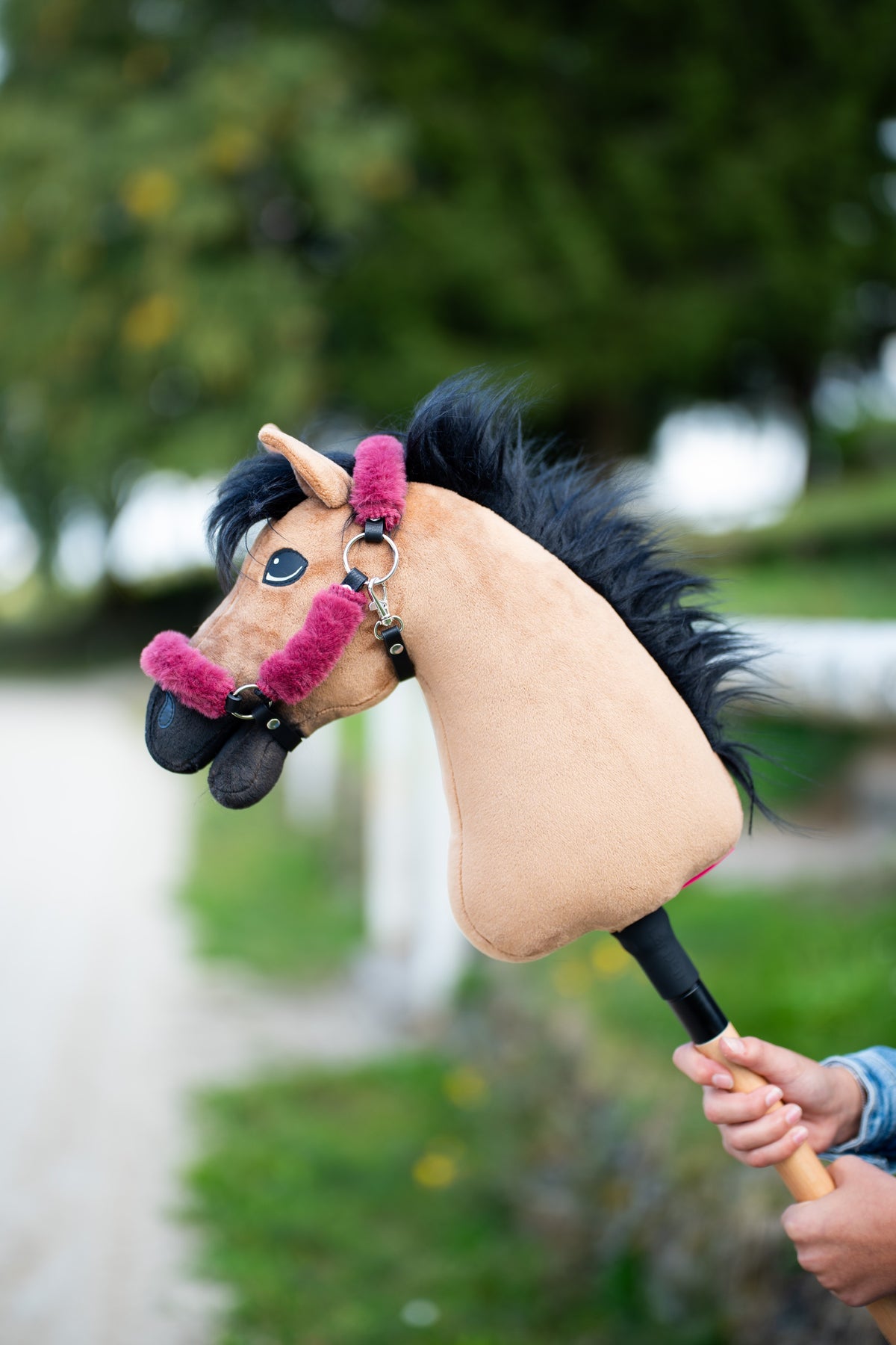 Detail photo: the head of the Hobby Horse Cookie with Black Halter by Hobby Horse LarDen held up beside a tree-lined track - sandy plush, long black yarn mane - shown here with the fur-lined Teddy Maroon halter.
