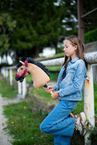 Lifestylefoto: een meisje in een spijkeroutfit leunt tegen een witte paddockreling en houdt de Hobby Horse Cookie met zwart Halster van Hobby Horse LarDen vast - zandkleurige pluchen kop, zwarte garenmane - hier getoond met het met bont gevoerde Teddy Maroon Halster