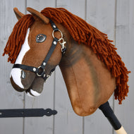 Hobby Horse Cooper met zwart halster van Hobby Horse LarDen, een bruine Hobby Horse geïnspireerd op een Oldenburger sportpaard. Dit speelgoed heeft een witte bles op het gezicht, dikke donkerbruine haren van garen en wordt geleverd met een zwart nylon halster met metalen fournituren. Bevestigd aan een houten stok en afgebeeld tegen grijze houten planken, ideaal als fantasierijk dressuurspel.