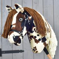 Hobby Horse Dakota met zwart halster van Hobby Horse LarDen, een pluchen model geïnspireerd op de mustang, met bruine manen van garen, zwart halster en een gevlekte bruin-witte vacht die de wilde geest van Noord-Amerika vastlegt, mooi getoond tegen een houten achtergrond.