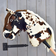 Hobby Horse Dakota met zwart halster van Hobby Horse LarDen, een pluchen stokpaard met bruine en witte vlekken, een hoofdstel en manen van garen geïnspireerd op de wilde Mustang-paarden van Noord-Amerika, getoond tegen een grijze houten wand.