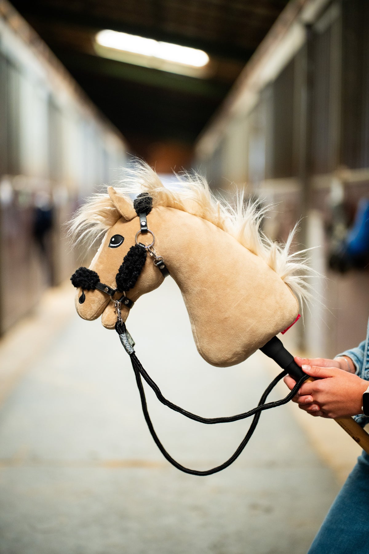 Detailfoto: de omhooggehouden kop van de Hobby Horse Dancing Lady met zwart Halster van Hobby Horse LarDen in een stalgang, met de Teugels naar beneden hangend - crèmekleurig pluche, lichte garenmane - hier getoond met het zwarte kunstleren Halster met volledig zwart bont en de zwarte Teugels