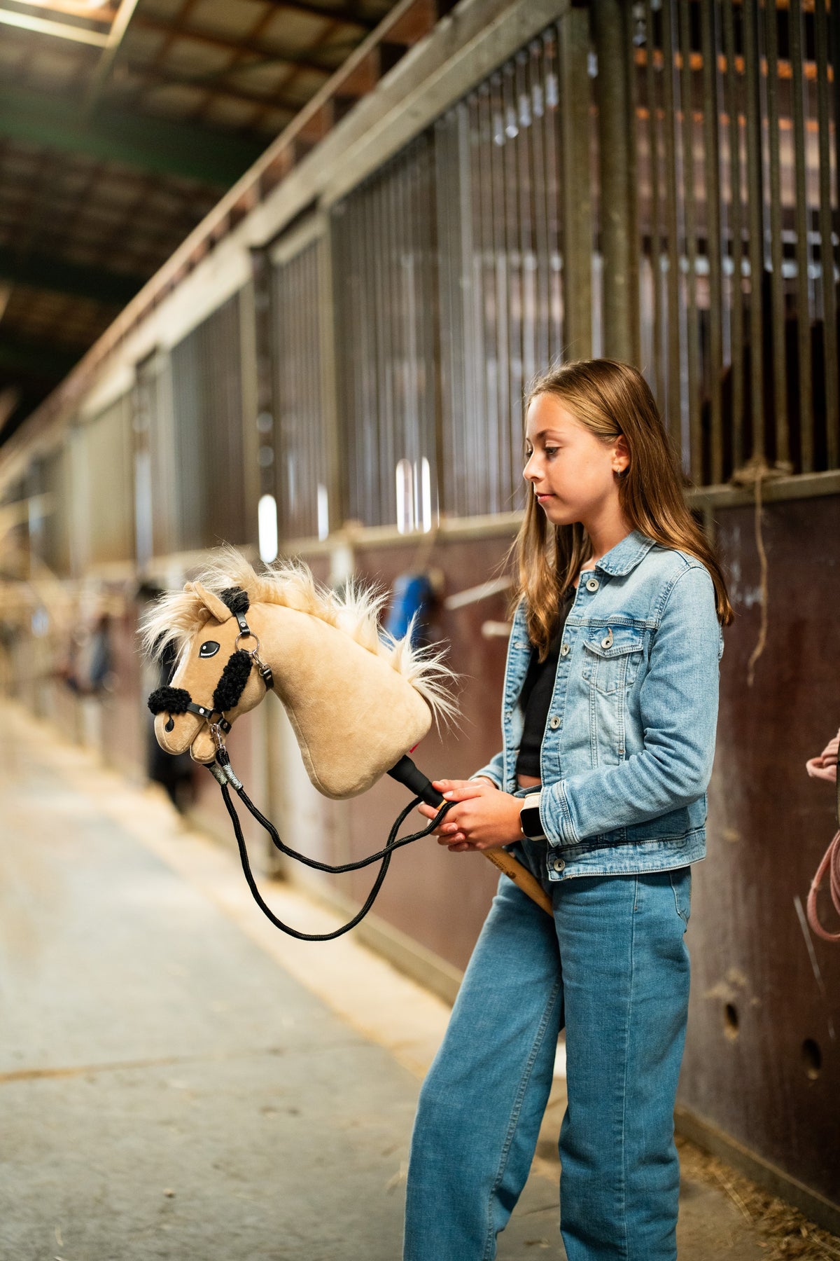 Lifestylefoto: een meisje in een spijkerjasje loopt met de Hobby Horse Dancing Lady met zwart Halster van Hobby Horse LarDen door een stalgang - crèmekleurige pluchen kop, lichte garenmane en voorpluk - hier getoond met het zwarte kunstleren Halster met volledig zwart bont en de zwarte Teugels