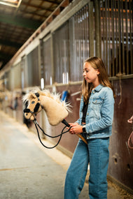 Lifestylefoto: een meisje in een spijkerjasje loopt met de Hobby Horse Dancing Lady met zwart Halster van Hobby Horse LarDen door een stalgang - crèmekleurige pluchen kop, lichte garenmane en voorpluk - hier getoond met het zwarte kunstleren Halster met volledig zwart bont en de zwarte Teugels