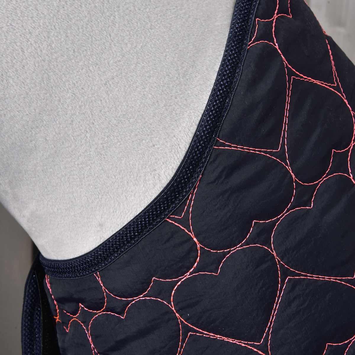 Close-up van Blanket Pink Heart van Hobby Horse LarDen, een donkere stoffen trainingsdeken voor paarden met roze hartvormige stikpatronen en donkerblauwe bies, gedrapeerd over een lichtgrijs oppervlak.