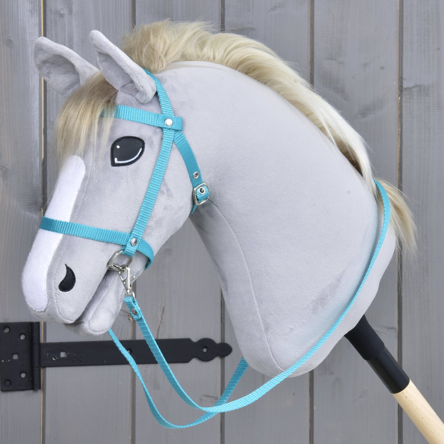 Grijze pluche Hobby Horse met lichte manen, met het Hobby Horse LarDen Zwart hoofdstel met teugels van turquoise band, gemonteerd op een houten stok en geplaatst voor een grijs houten hek.