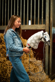 Lifestylefoto: een meisje in een spijkerjasje staat voor strobalen in een stal en houdt de Hobby Horse Elektra met zwart Halster van Hobby Horse LarDen vast - wit gevlekt pluchen kop, blauw oog, witte garenmane - hier getoond met het Graphite Full bonthalster van kunstleer en het gebreide Halstertouw