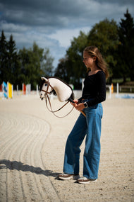 Lifestylefoto: een meisje in een zwarte top staat op een zandpiste onder een stormachtige hemel en houdt de Hobby Horse Frederik LD met zwart Halster van Hobby Horse LarDen vast, met gekleurde springbalken erachter - crèmekleurige pluchen kop, donkerbruine garenmane - hier getoond met het volledige bruine hoofdstel met Neusriem