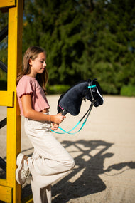 Lifestylefoto: een meisje in een roze top leunt tegen een gele hindernisstaander en houdt de Hobby Horse Freya met zwart Halster van Hobby Horse LarDen vast - zwarte pluchen kop, witte bles, zwarte garenmane - hier getoond met het volledige turquoise hoofdstel zonder Neusriem