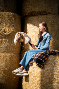 Lifestylefoto: een meisje in een spijkerjasje zit hoog op ronde strobalen en houdt de Hobby Horse Fury met zwart Halster van Hobby Horse LarDen vast - palomino pluchen kop met witte aftekeningen, crèmekleurige garenmane - hier getoond met het met bont gevoerde LarDen Brown Halster en het Bloom Brown Halstertouw