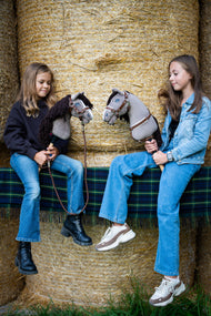 Twee meisjes zitten op strobalen op een geruite deken, elk met een Hobby Horse Guccini van Hobby Horse LarDen met een grijze kop in Holsteiner-stijl, witte neusstreep en lange donkerbruine garenmanen, met het met bont gevoerde Halster Walnut, het borsttuig Walnut met bont en het Halstertouw Vivaldi