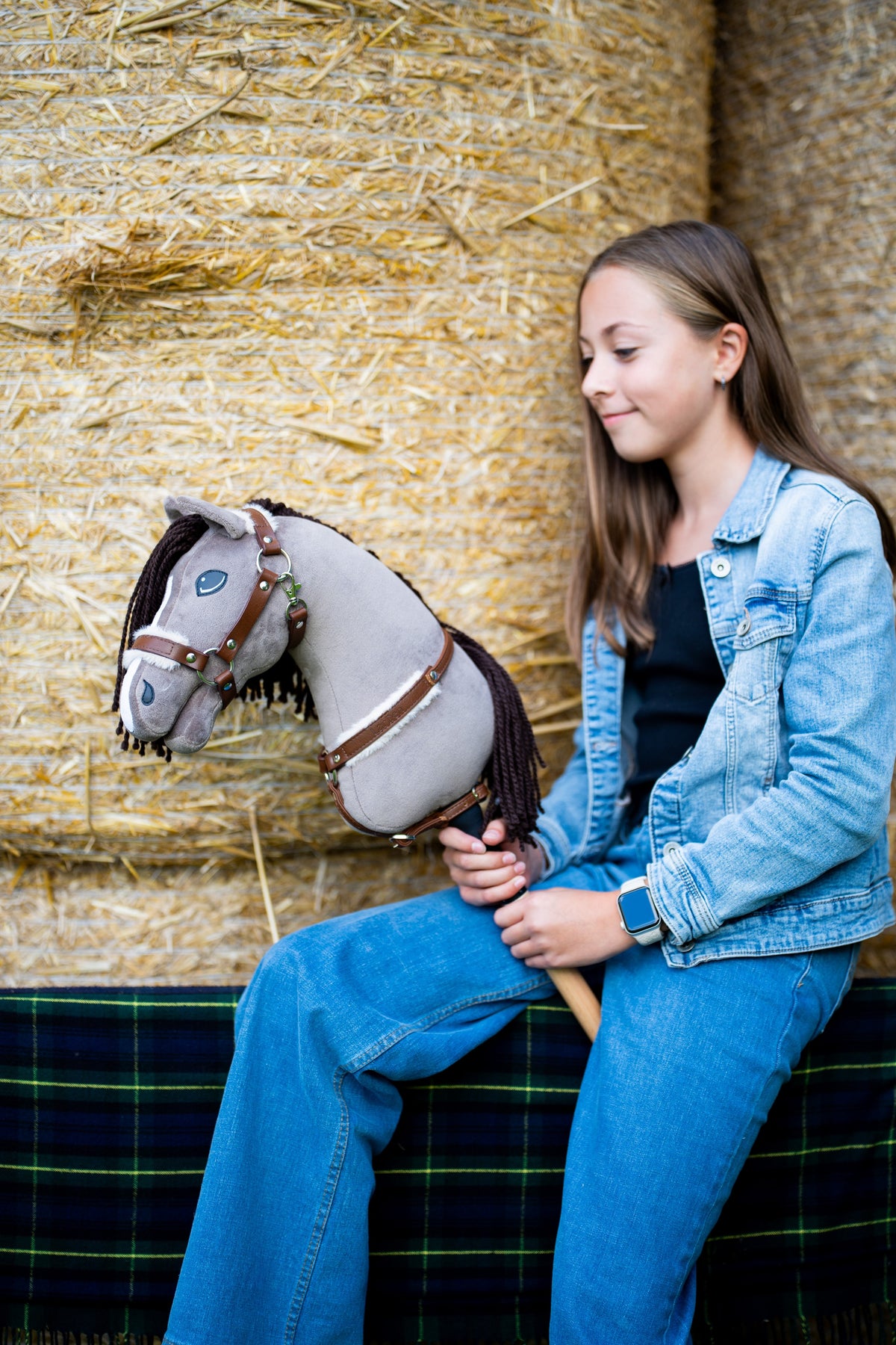 Lifestyle photo: a girl in a denim jacket sits on straw bales holding the Hobby Horse Guccini by Hobby Horse LarDen - grey head, white nose stripe, long dark brown yarn mane, wooden stick - shown here with the fur-lined Walnut halter and the Walnut breastgirth with fur.