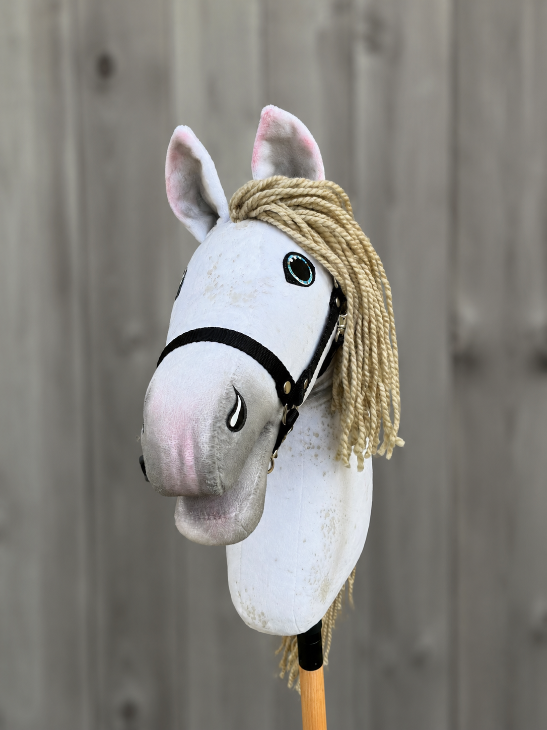 The Hobby Horse LarDen "Victory" (size M) is a white hobby horse inspired by the Andalusian, featuring a black halter and blond yarn mane on a wooden stick, shown against a blurred wooden fence background.