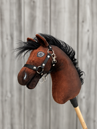 Hobby Horse Benny met Zwart Halster - maat XS van Hobby Horse LarDen, een bruine pluchen Hobby Horse met zwarte manen, met een zwart halster, op een houten stok voor een onscherpe achtergrond van een houten hek.