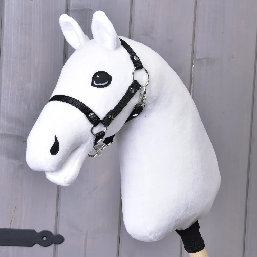 Bouw je paard, aangepast wit Hobby Horse met opvallende zwarte manen, vrolijke uitdrukking, zwart hoofdstel en houten stok, weergegeven voor een lichtgrijze houten wand.