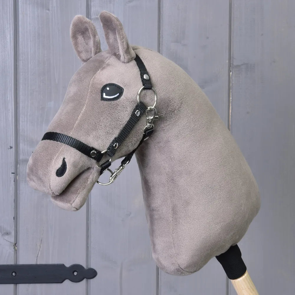 Grijze pluchen Hobby Horse-kop met een zwart hoofdstel, gemonteerd op een houten stok en getoond voor een houten wand, ideaal als accessoire voor de Bouw je paard-kit voor het personaliseren van je Hobby Horse voor fantasierijk spel.
