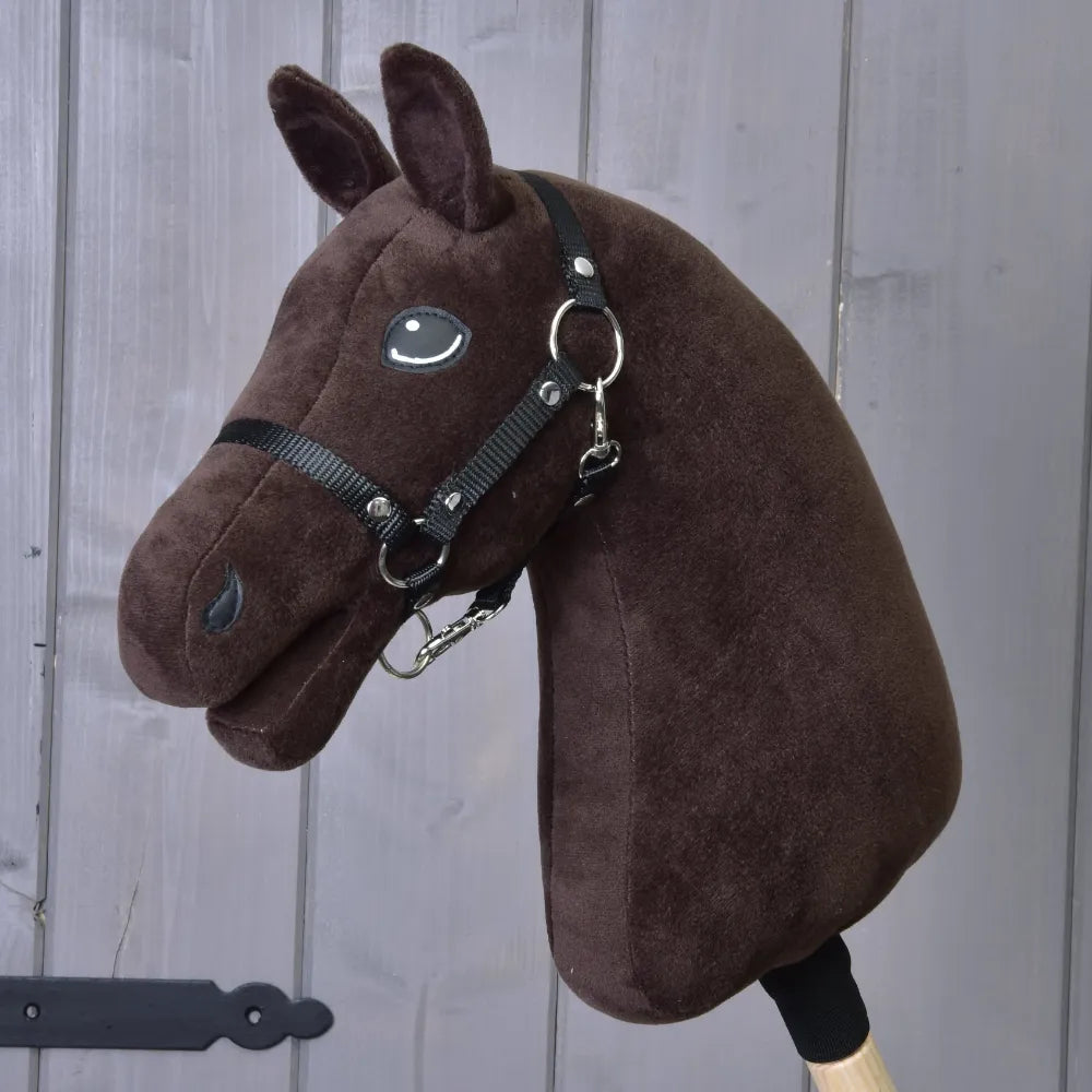 Bouw je paard, aanpasbare bruine pluchen Hobby Horse-kop met zwart hoofdstel en teugels, gemonteerd op een houten stok en weergegeven tegen een grijze houten muur.