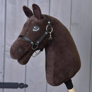 Bouw je paard, aanpasbare bruine pluchen Hobby Horse-kop met zwart hoofdstel en teugels, gemonteerd op een houten stok en weergegeven tegen een grijze houten muur.