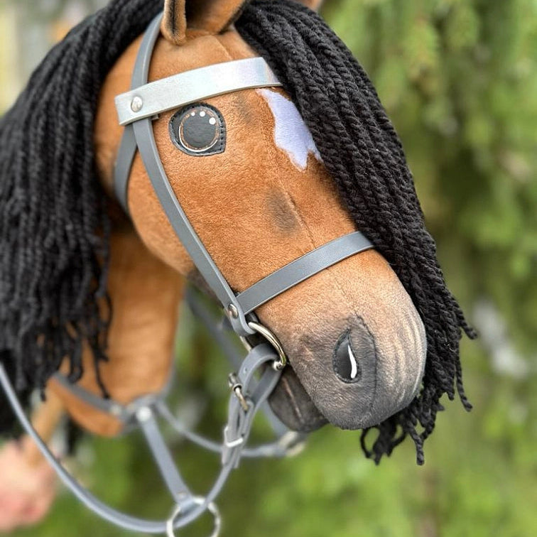 Close-up van een handgemaakte Hobby Horse met een zwarte manen van garen, bruine vacht, een witte vlek boven één oog en een grijs hoofdstel, tegen een onscherpe groene buitenachtergrond, een perfect accessoire voor Hobby Horsing-avonturen.